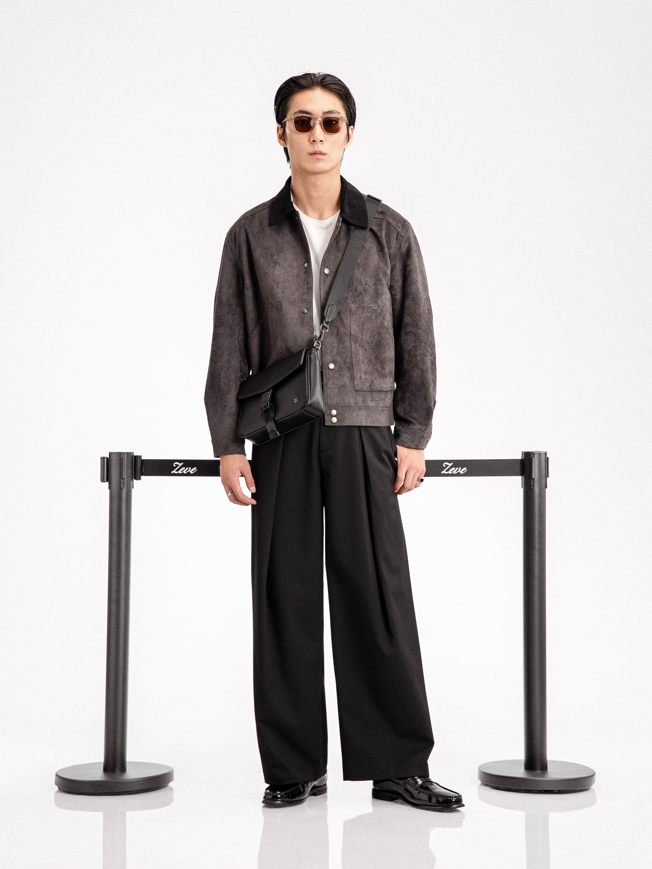 Man in stylish black loafers, wide-leg pants, and suede jacket with crossbody bag, sunglasses, minimalist studio setting