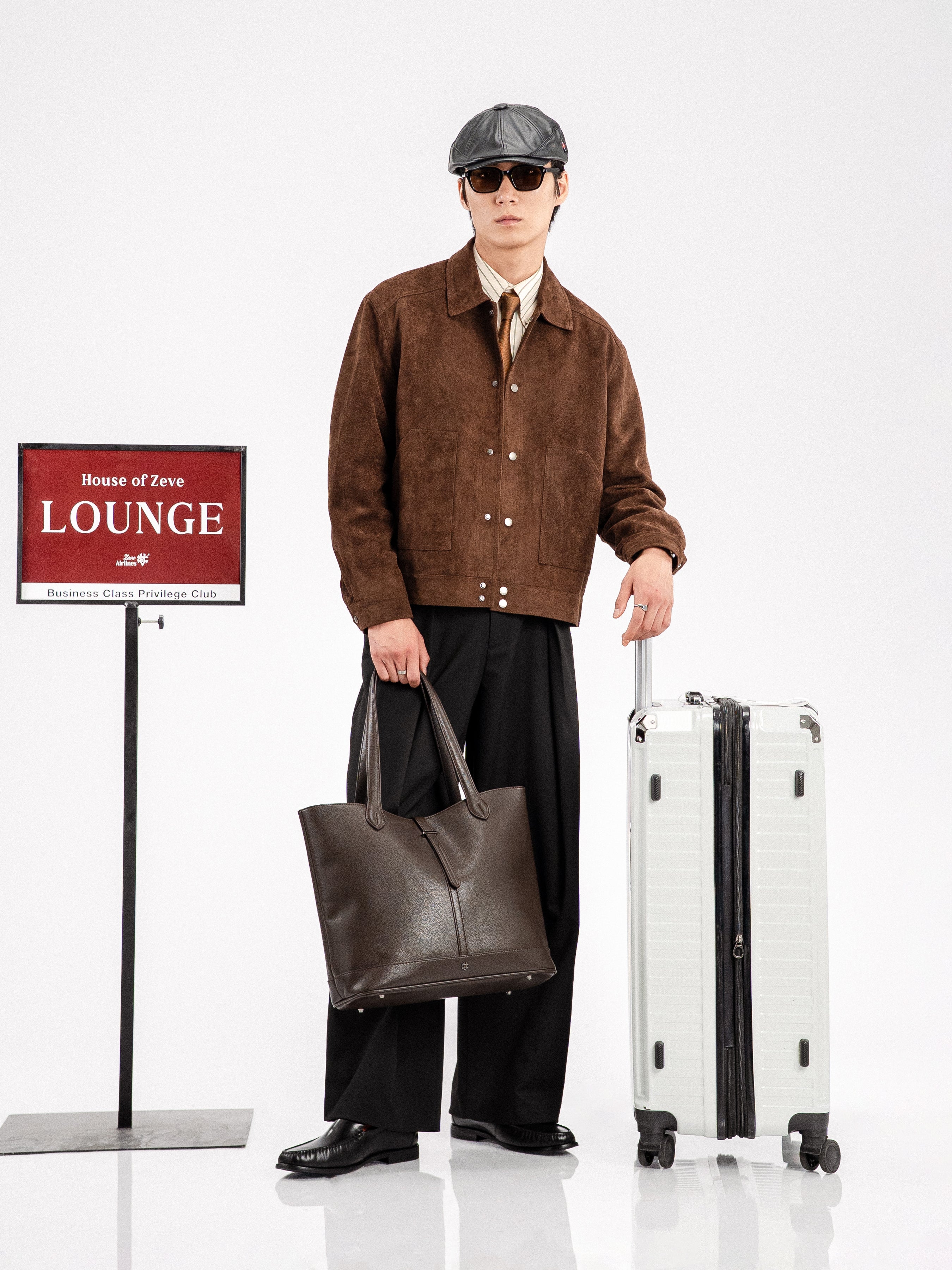 Man in brown suede jacket, black loafers, holding leather tote and suitcase near lounge sign