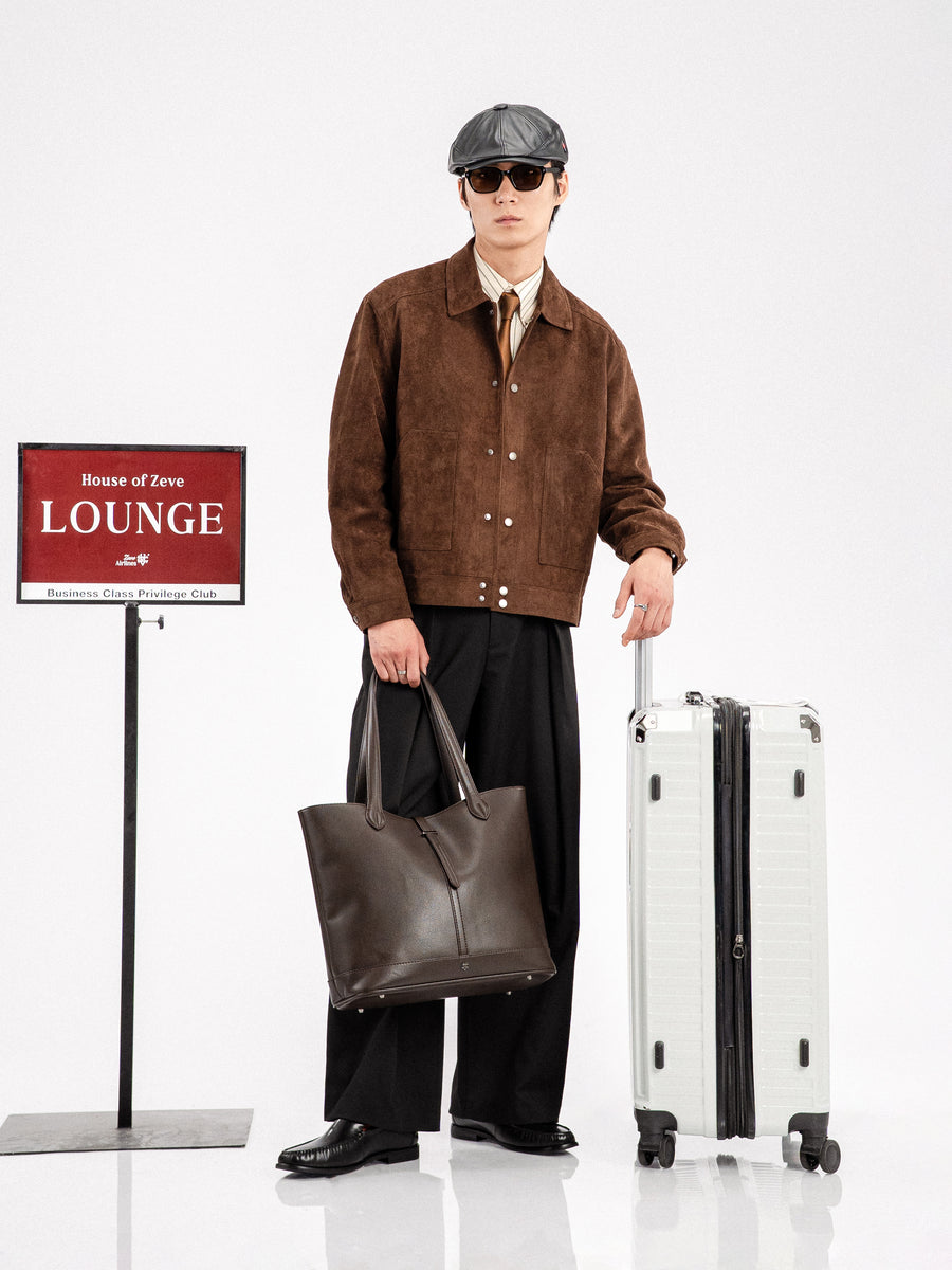 Man in brown suede jacket, black loafers, holding leather tote and suitcase near lounge sign